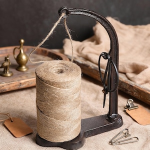 May include: A rustic, dark metal twine holder with a spool of natural-colored twine. The holder has a curved arm and a base, with a pair of black scissors hanging from it. A small brown tag and safety pins are also visible.