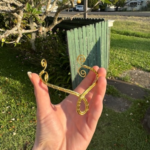 May include: A gold-coloured wire headpiece with a decorative, swirling design. The headpiece is held in a hand against a background of green grass and a green fence.