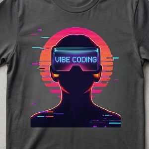 May include: Dark gray t-shirt featuring a neon graphic of a person wearing VR goggles with the text "VIBE CODING". The design includes a retro sunset and colorful accents. The shirt is surrounded by accessories like a notebook, headphones, and succulents.