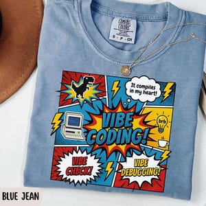 May include: A blue jean colored t-shirt featuring a comic book style graphic with the text VIBE CODING, It compiles in my heart, VIBE CHECK, and VIBE DEBUGGING. The design includes a pixelated dinosaur, a computer, a lightbulb, and lightning bolts.