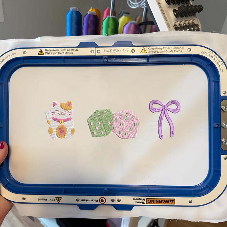 May include: A blue rectangular embroidery hoop holding white fabric with three embroidered designs: a white and yellow beckoning cat, a pair of green and pink dice, and a purple ribbon bow. The hoop frame includes text labels and warning icons.