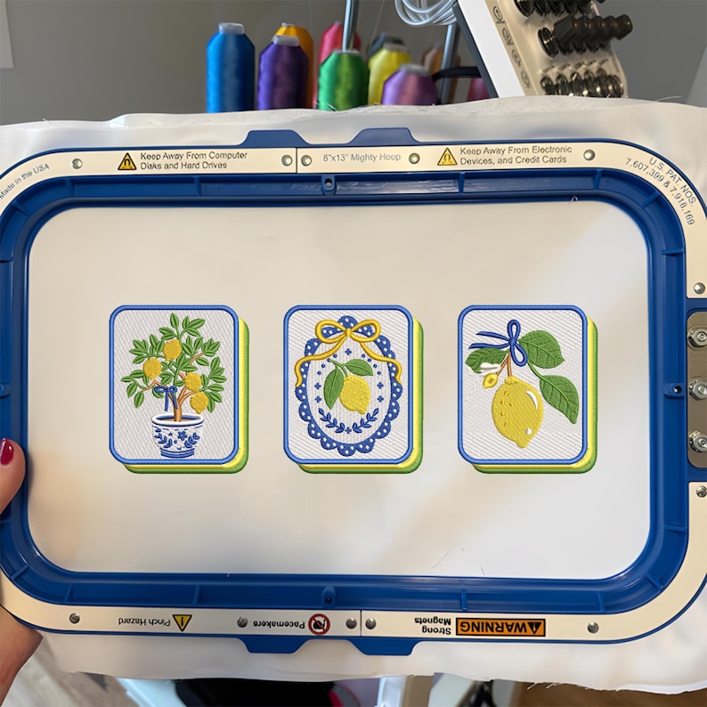 May include: A blue 8x13 inch embroidery hoop holding white fabric with three embroidered lemon designs. The left shows a lemon tree in a blue and white pot, the center a lemon in a blue oval frame with a yellow bow, and the right a lemon branch with a blue bow.