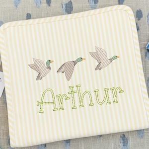 Könnte beinhalten: Eine quadratische Babydecke mit gelb-weißem Streifenmuster. Sie zeigt drei gestickte Enten und den Namen "Arthur" in grüner Schrift. Die Decke hat einen weißen Rand.