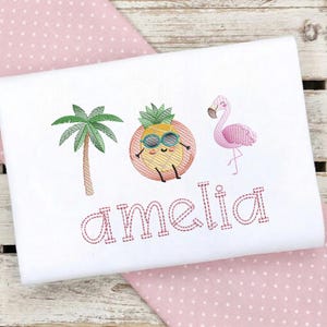Puede incluir: Camiseta blanca con diseños tropicales bordados. Presenta una palmera, una piña sonriente con gafas de sol y un flamenco rosa. El nombre "amelia" está bordado en rosa debajo.