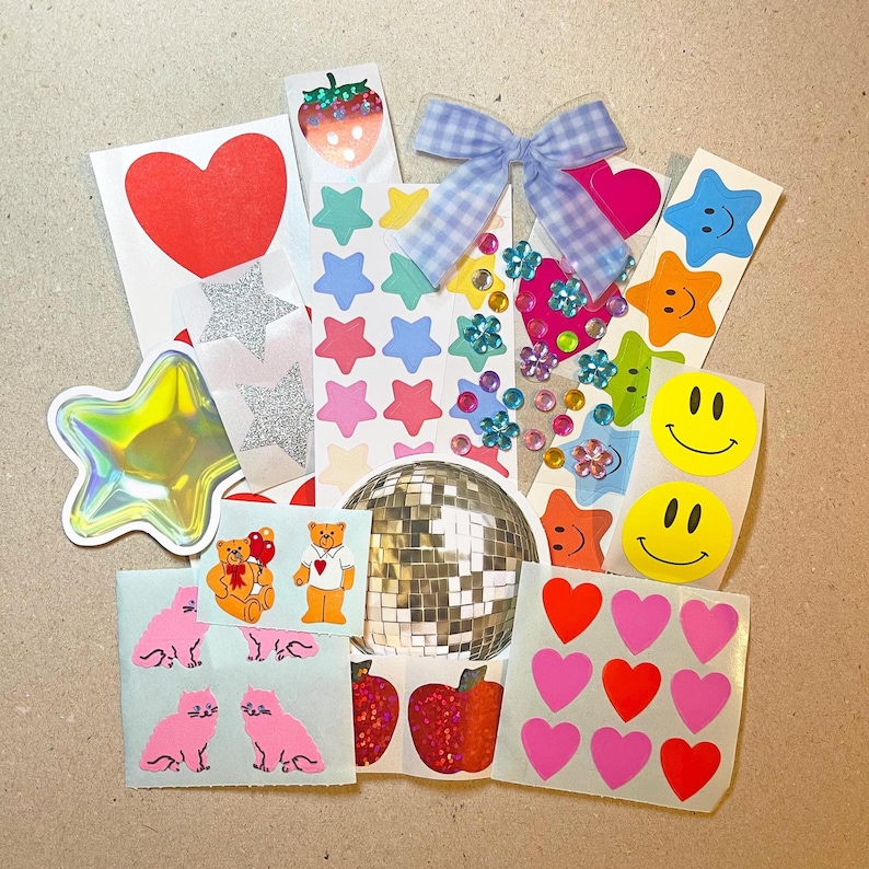 May include: A collection of colorful stickers featuring various designs. Includes a red heart, glitter stars, smiley faces, star shapes, a disco ball, and a gingham bow. Also includes stickers of cats, bears, and strawberries.