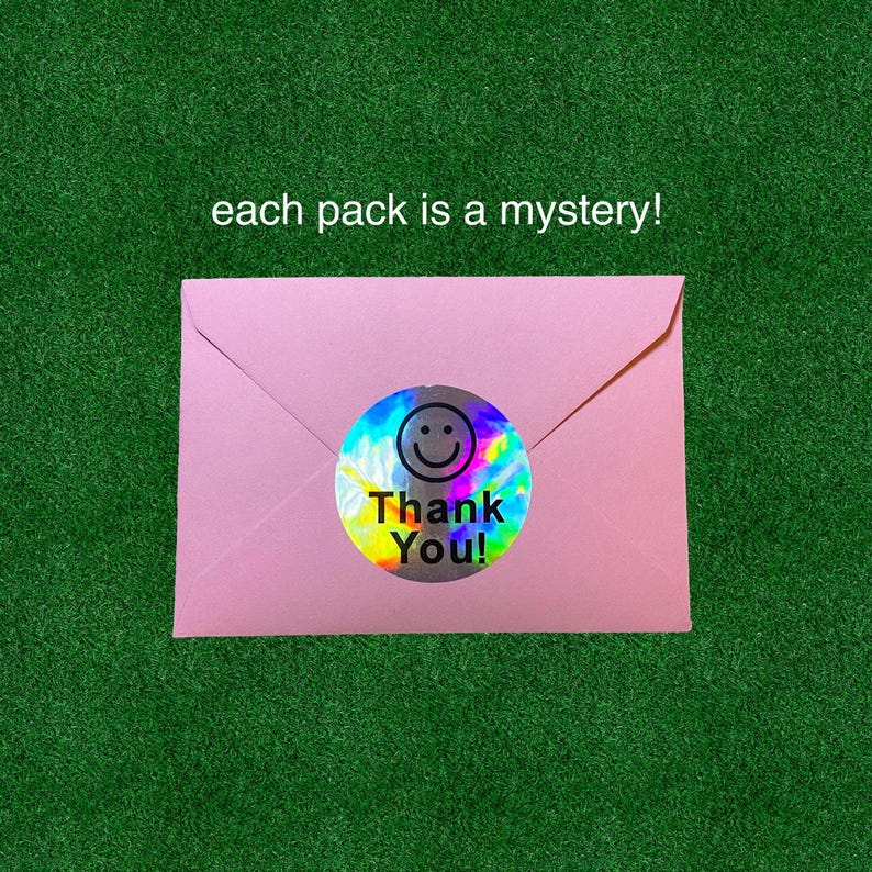 May include: A pink envelope sealed with a holographic sticker that says "Thank You!" and features a smiley face. The text "each pack is a mystery!" is above the envelope. The background is green.