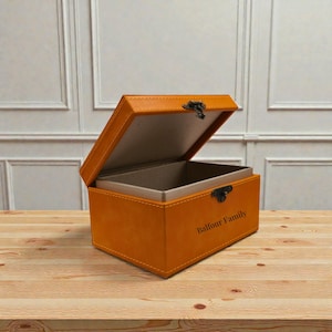 May include: An open, orange leather box with a hinged lid and a metal clasp. The box has a lighter-colored interior and the words "Balfour Family" are printed on the front. The box is sitting on a wooden surface.