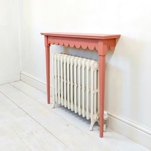 May include: A coral-coloured wooden radiator cover console table featuring a scalloped edge design along the front apron, supported by four straight legs, positioned over a white cast iron radiator with vertical columns and a side valve.