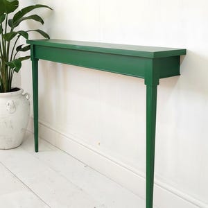 May include: A narrow, rectangular console table finished in a solid deep green paint. The table features a flat top surface, a straight apron, and four slender, tapered legs that extend vertically to the floor, providing a stable base.