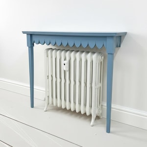 May include: A blue scalloped-edge console table with a white radiator beneath it. The table has a simple design with four legs and a light blue finish. The radiator is a classic cast iron model.