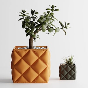 Op de afbeelding: Twee vierkante plantenbakken met een gewatteerd ontwerp. De grotere, oranje plantenbak bevat een jadeplant. De kleinere, donkergroene plantenbak bevat een vetplant. Beide staan op een witte achtergrond. De grotere bak is ongeveer 25 cm breed.