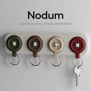 May include: A wall-mounted key holder labelled Nodum with the text Spinning cores. Fixed destination. It features four circular button-shaped hooks in olive green, brown, beige, and red, each with a white cross-stitch detail and a silver key ring.