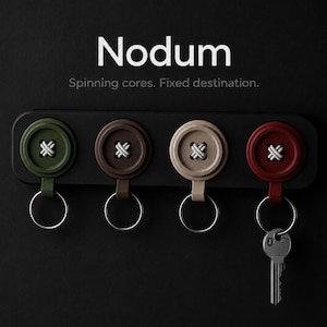 May include: A black wall-mounted key holder featuring four circular button-shaped hooks in olive green, brown, beige, and red. Each button has a white cross-stitch detail and a silver metal ring. The text reads: Nodum. Spinning cores. Fixed destination.