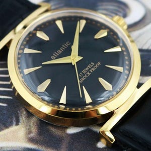 May include: Close-up of a gold-toned wristwatch with a black face. The watch features gold hands and markers, the brand name "atlantic", and the text "17 JEWELS SHOCK PROOF". The watch has a black leather band.