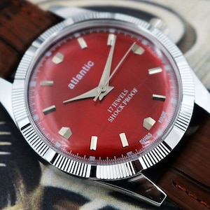 Puede incluir: Primer plano de un reloj de pulsera Atlantic vintage con esfera roja vibrante. El reloj presenta agujas y marcadores plateados, un bisel plateado y una correa de cuero marrón. La esfera del reloj indica "Atlantic", "17 Jewels" y "Shock Proof".