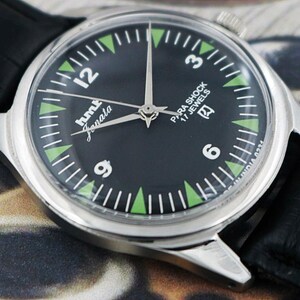 May include: A close-up of a silver-toned wristwatch with a black face. The watch features white numerals, green triangular markers, and the text "hmt Janata" and "Para Shock 17 Jewels". The watch has a black leather band.
