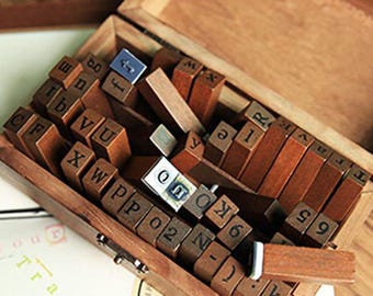 May include: A wooden box filled with individual letter and number stamps. The stamps are arranged inside a wooden box. The box is open, revealing the stamps, and is sitting on top of a passport and a wooden surface.