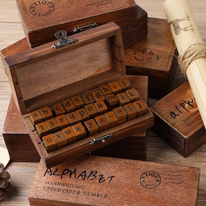 May include: Wooden alphabet stamp sets with uppercase and lowercase letters, numbers, and symbols. The stamps are stored in small, antique-style wooden boxes. One box is open, revealing the letter stamps. The boxes are brown with black text.
