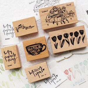 May include: A collection of wooden stamps with various designs and text. Designs include musical notes, a group of people, tulips, a coffee cup with the text "Start Your Day with Coffee", and the words "Missing You". The stamps are on a white surface.
