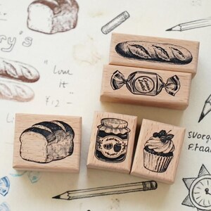 Könnte beinhalten: Fünf rechteckige Holzstempel mit schwarzen Tintenillustrationen eines Baguettes, eines verpackten Bonbons, eines Brotlaibs, eines Marmeladenglases und eines Cupcakes. Die Stempel liegen auf einer Seite mit verschiedenen Skizzen und Text.