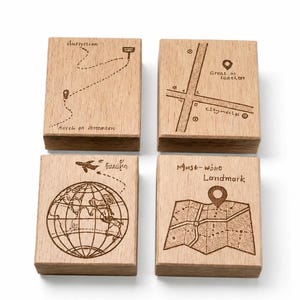 Könnte beinhalten: Vier Holzblöcke mit gravierten Designs zum Thema Reisen. Ein Block zeigt eine gepunktete Linie und ein Zielsymbol. Ein anderer zeigt einen Stadtplan mit einer Standortmarkierung. Der dritte Block hat einen Globus und ein Flugzeug. Der letzte Block zeigt eine Karte mit einem Landmarken-Pin.