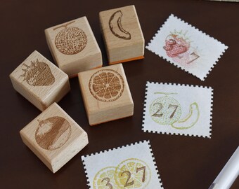 May include: Five square wooden stamps with engraved fruit designs including a melon, banana, strawberry, orange slice, and chestnut. Three rectangular white paper labels feature stamped fruit illustrations and the numbers 3, 7, and 27 in brown ink.