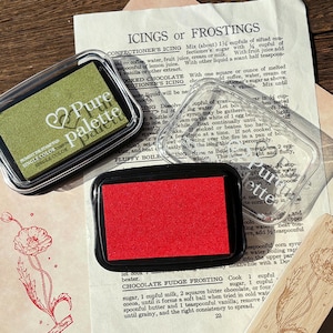 May include: Two ink stamp pads with clear lids, one green and one red, sit on a vintage recipe page. The green pad is labeled "Pure Palette" and "Single Color". The red pad is next to a stamped floral design.