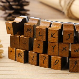 May include: Wooden alphabet stamps with black letter and number imprints. The stamps are arranged in a stack, showing letters like N, M, L, K, J, and numbers like 0, 7, and 9. The stamps are a warm brown color.