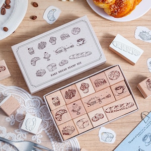 May include: A rectangular cardboard box labelled BAKE BREAD STAMP SET containing twelve wooden stamps with engraved illustrations of various baked goods, including a croissant, a baguette, a cupcake, a coffee cup, and a fork with the text sweet.