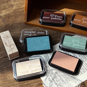 May include: An assortment of ink pads in various colors, including blue, brown, and peach, with the text "Pure palette" on the lids. A wooden stamp with a floral design is also present. The ink pads are in clear plastic cases.