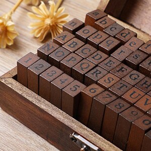 May include: A wooden stamp set with letters and numbers arranged in a wooden box. The stamps are dark brown with black lettering. The box is open, revealing the stamps in neat rows. The set includes the alphabet and numbers 1-9.