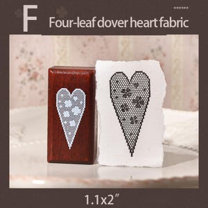 May include: A rectangular wooden stamp with a white lace-style heart pattern featuring four-leaf clovers, next to a piece of textured white paper showing a black stamped impression of the same heart design. The text F Four-leaf dover heart fabric 1.1x2" is visible.