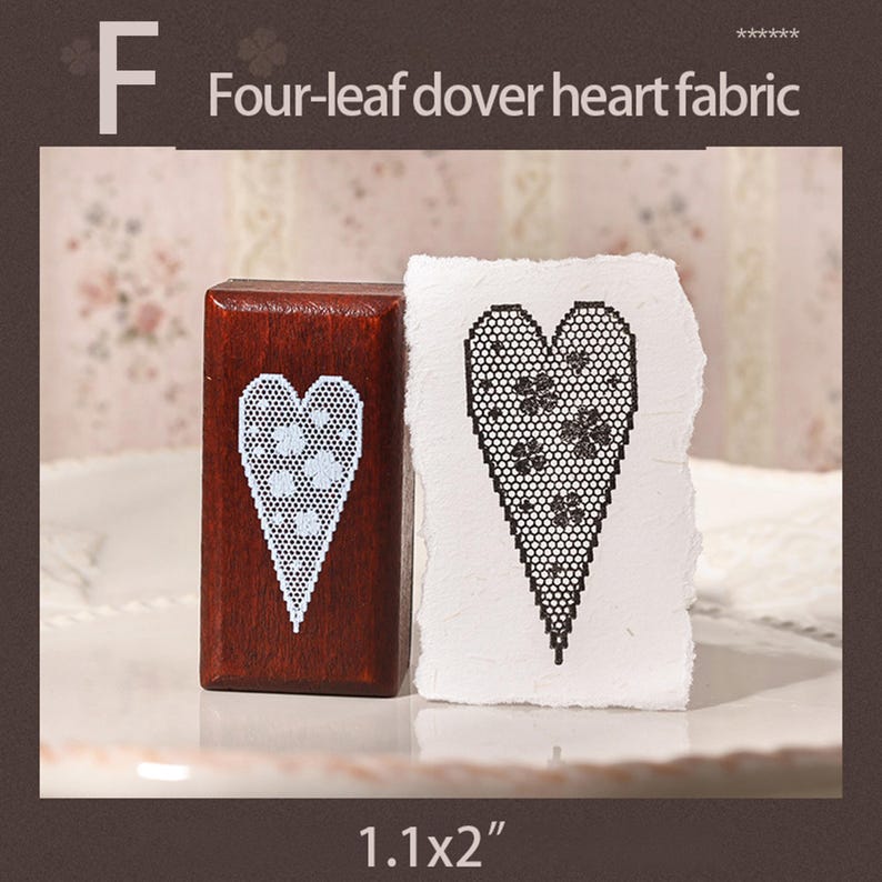 May include: A rectangular wooden stamp with a white lace-style heart pattern featuring four-leaf clovers, next to a piece of textured white paper showing a black stamped impression of the same heart design. The text F Four-leaf dover heart fabric 1.1x2" is visible.