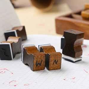 May include: Wooden letter stamps with dark brown wooden handles and black ink pads. The letters "W" and "N" are visible on two of the stamps. The stamps are on a white grid paper.