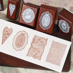 May include: A collection of four wooden rubber stamps with dark brown blocks, each featuring a white lace-like design on top. A white paper strip displays four stamped impressions in a reddish-brown ink, showing a heart, an oval, and two rectangular lace patterns.