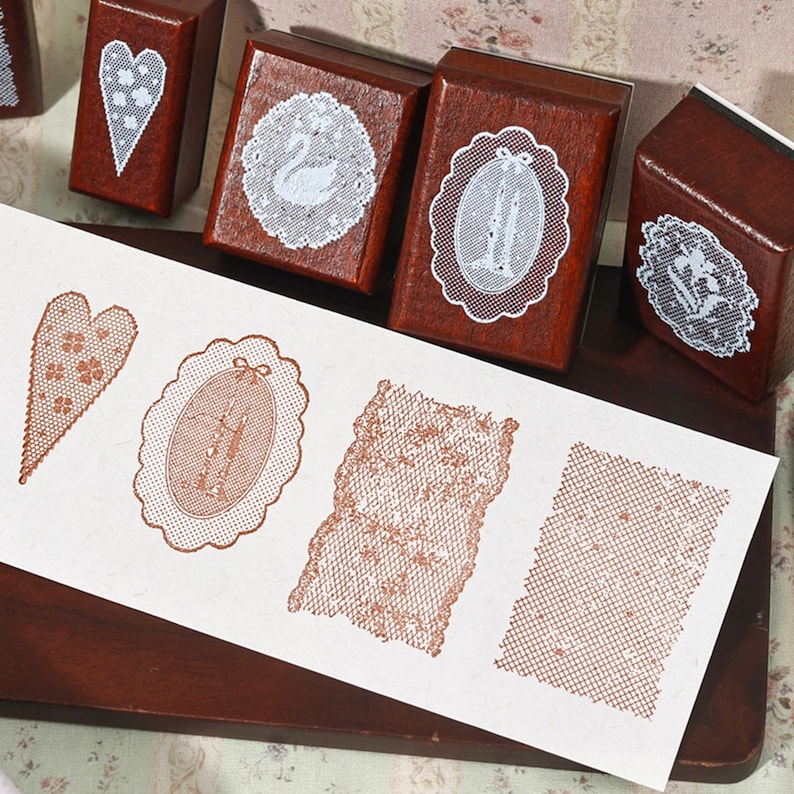 May include: A collection of four wooden rubber stamps with dark brown blocks, each featuring a white lace-like design on top. A white paper strip displays four stamped impressions in a reddish-brown ink, showing a heart, an oval, and two rectangular lace patterns.