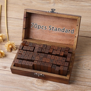May include: A wooden box with a hinged lid, containing a set of 70 standard wooden letter stamps. The stamps are arranged in rows, with both uppercase and lowercase letters, numbers, and symbols. The box is made of dark brown wood.