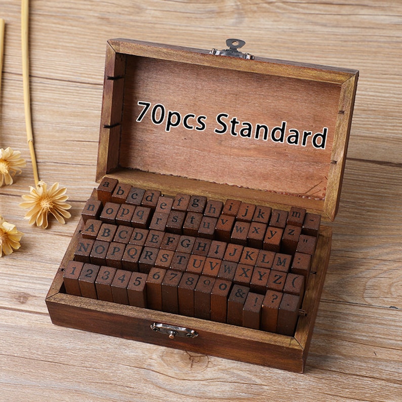 May include: A wooden box with a hinged lid, containing a set of 70 standard wooden letter stamps. The stamps are arranged in rows, with both uppercase and lowercase letters, numbers, and symbols. The box is made of dark brown wood.