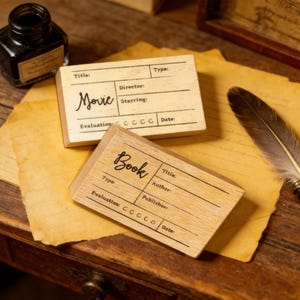 Könnte beinhalten: Zwei Holzstempel, einer mit der Aufschrift "Movie" und der andere mit der Aufschrift "Book", mit Feldern für Titel, Regisseur, Autor und andere Details. Die Stempel liegen auf einer Holzoberfläche mit gealtertem Papier und einer Feder.
