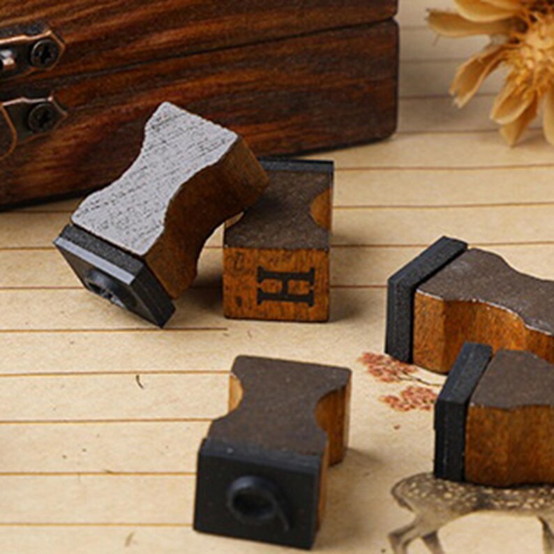 May include: A collection of vintage wooden letter stamps with black rubber printing surfaces. The stamps have a natural wood grain finish and are arranged on a patterned surface. One stamp displays the letter "H".