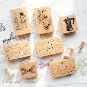 May include: A collection of wooden stamps with various designs. The stamps feature images of an astronaut, a swan, a coffee pot, stars, and a bow. The stamps are made of light-colored wood and have black ink designs. The words "Wood seal" are at the bottom.