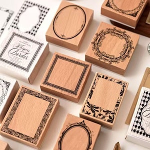 Puede incluir: Una colección de sellos de madera con varios diseños de bordes decorativos. Algunos sellos presentan marcos ovalados, cuadrados y florales. Un sello dice "Whispers in the Border". Los sellos están dispuestos sobre una superficie blanca.