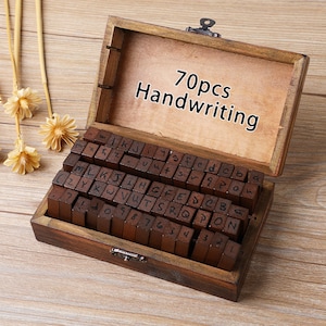 May include: A wooden box containing 70 handwriting stamps. The stamps are arranged in rows and columns inside the box. The box is open, revealing the stamps and the text "70pcs Handwriting" on the inside of the lid. The box is made of dark wood.