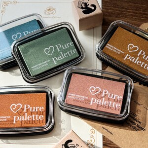 May include: An assortment of pigment ink stamp pads in various colors, including teal, green, and orange, with the text "Pure palette" and a heart symbol. Each pad is in a clear plastic case. A wooden stamp with a cat illustration is also visible.