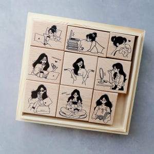 May include: A set of nine square wooden stamps arranged in a light wood tray. Each stamp features a black line art illustration of a person in various daily activities, including using a laptop, applying makeup, holding a pet, and playing games.
