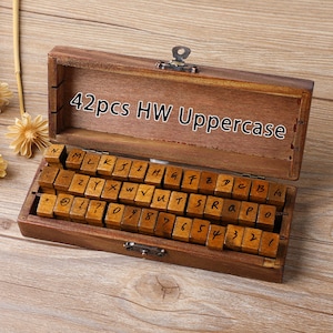 May include: A wooden box containing 42 uppercase letter stamps and numbers. The stamps are arranged in rows inside the open box. The box is made of dark brown wood and has a metal clasp.