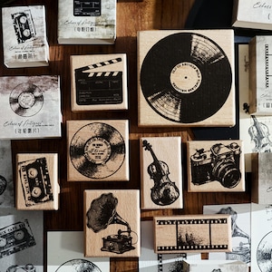 May include: A collection of square and rectangular wooden rubber stamps featuring black ink illustrations of vintage items including a vinyl record, a film clapperboard, a cassette tape, a violin, a camera, a gramophone, and a strip of film.