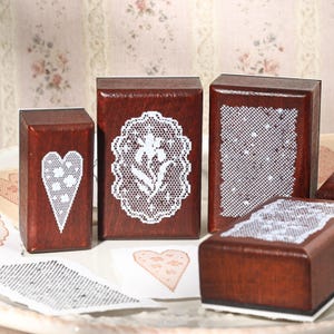 May include: Four rectangular wooden stamps with dark brown finish. One features a white lace heart, one a white lace floral oval, one a white lace grid pattern, and one a white lace border design. Stamped paper samples are visible below.