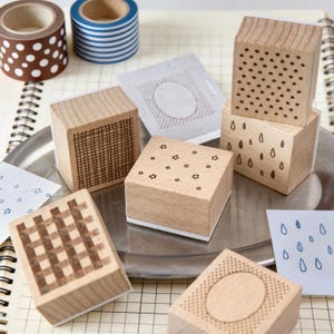 May include: A collection of six square wooden stamps featuring various engraved patterns including a grid, stars, raindrops, dots, and an oval frame, accompanied by two rolls of patterned washi tape and several stamped paper samples.
