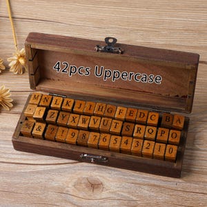 May include: A wooden box containing 42 uppercase letter stamps and numbers. The stamps are arranged in rows inside the open box. The box is made of dark brown wood and has a metal clasp. The text "42pcs Uppercase" is printed on the inside of the lid.